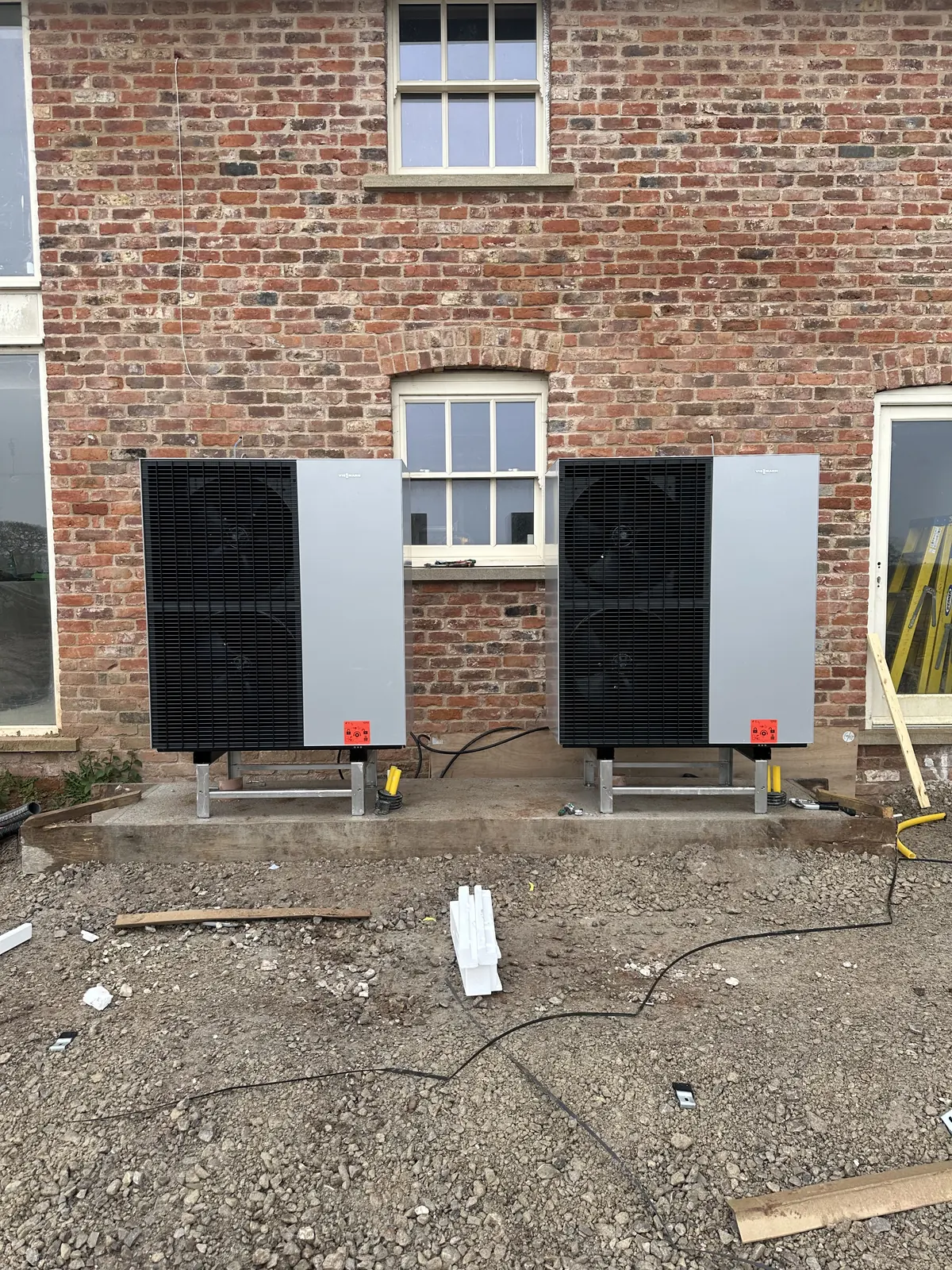 Two air source heat pumps on frames against a brick wall